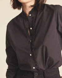 A woman with chin-length hair, partially out of frame, wears the Trovata Birds of Paradis Helena Shirt in Black—a cotton blouse with a ruffle collar and placket—paired with black pants.