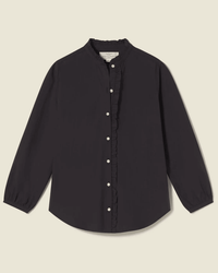 The Helena Shirt in Black by Trovata Birds of Paradis is a women’s cotton blouse with a ruffle collar, button-down front with white buttons, ruffled trim, and gathered cuffs, shown against a simple light background.