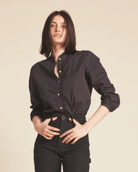 A person with straight, shoulder-length dark hair wears the Trovata Birds of Paradis Helena Shirt in Black and black pants, standing against a plain beige background with a relaxed expression and hands on their waist.