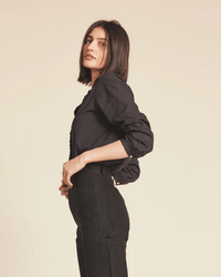 A woman with straight, shoulder-length dark hair wears the Trovata Birds of Paradis Helena Shirt in Black, paired with high-waisted pants. She stands sideways against a plain beige background, looking toward the camera.