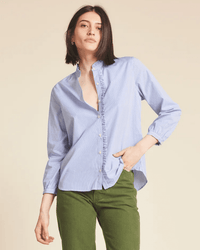 A person with straight, shoulder-length dark hair wears the Helena Shirt in Blue/White Stripe by Trovata Birds of Paradis, a ruffle collar button-down, partially unbuttoned and paired with green pants against a plain beige background.