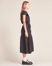 A woman in profile stands against a neutral background, wearing the Kristi Dress in Black by Trovata Birds of Paradis, paired with white socks and black loafers. Her long hair is loose and she gazes straight ahead.