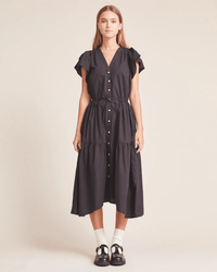 A woman faces forward wearing the Kristi Dress in Black by Trovata Birds of Paradis, featuring buttons down the front, ruffle sleeves, and a drawstring waist. She completes the look with white socks and black shoes against a beige background.