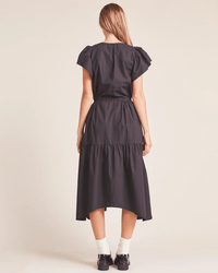 A woman with long light brown hair faces away, wearing the Trovata Birds of Paradis Kristi Dress in Black—a midi dress with ruffle sleeves and a tiered skirt—paired with white socks and black shoes against a plain light background.