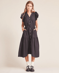 A woman faces forward in the Kristi Dress in Black by Trovata Birds of Paradis, featuring ruffle sleeves and a cinched waist, styled with white socks and black shoes against a plain beige background.
