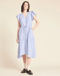 A woman stands against a beige background wearing the Trovata Birds of Paradis Kristi Dress in Blue/White Stripe with ruffle sleeves. She pairs it with white socks and black loafers, one hand in her pocket and a relaxed expression.