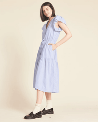 A person stands sideways in the Kristi Dress in Blue/White Stripe by Trovata Birds of Paradis, featuring short ruffle sleeves and side pockets. They wear white socks and black loafers against a plain beige background.