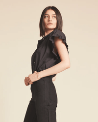 A woman with straight dark hair models the "Marianne B in Black" top by Trovata Birds of Paradis, standing sideways and glancing over her shoulder against a beige background.