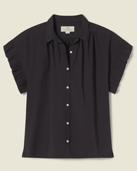 The Marianne B in Black by Trovata Birds of Paradis is a short-sleeve ruffle-collared shirt with white buttons, shown on a light beige background.