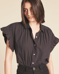 A woman with shoulder-length dark hair wears the Marianne B in Black shirt by Trovata Birds of Paradis, featuring loose-fit ruffle sleeves and white buttons, paired with high-waisted black pants against a neutral background.