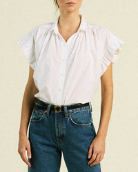 A woman wears the Marianne B Ruffle Sleeve Shirt in White by Trovata Birds of Paradis, tucked into high-waisted blue jeans with a black belt and gold buckle, standing against a plain light background.