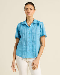 A woman wearing the Trovata Birds of Paradis Millie Blouse in Larkspur, a short-sleeved light blue pintuck button down with pleated details, pairs it with white pants against a plain beige background.