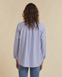 A person with wavy, shoulder-length hair is seen from behind, wearing a blue/white striped Sara B Henley Shirt by Trovata Birds of Paradis and blue jeans, standing against a beige background.
