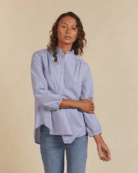 A person wears a Trovata Birds of Paradis Sara B Henley Shirt in Blue/White Stripe with blue jeans, standing against a plain beige background.