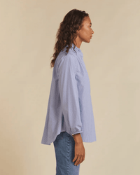 A woman stands in profile against a beige background, wearing a loose-fitting Sara B Henley Shirt in Blue/White Stripe by Trovata Birds of Paradis and blue jeans.