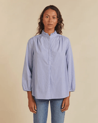 A person wearing the Trovata Birds of Paradis Sara B Henley Shirt in Blue/White Stripe with light blue jeans stands against a plain beige background.