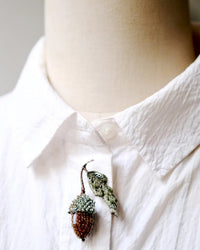 A close-up of a person in a textured white collared shirt, featuring the Trovelore Acorn Brooch Pin—hand embroidered and acorn-shaped—pinned near the top button.