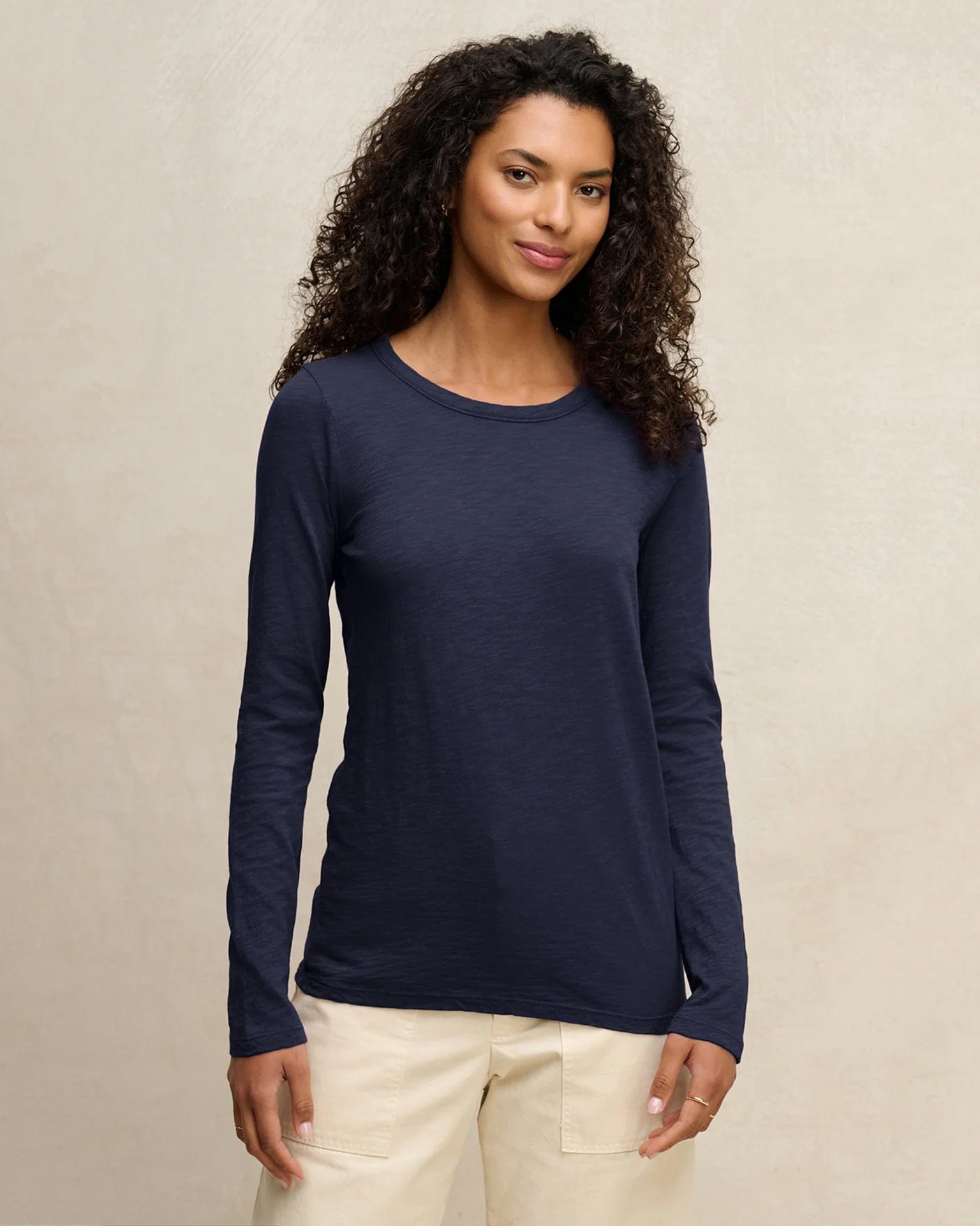 A woman with curly hair wears the Lizzie Long Sleeve Crew in Midnight by Velvet by Graham & Spencer with cream pants, standing against a neutral background and smiling softly at the camera.