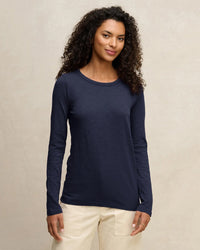 A woman with curly hair wears the Lizzie Long Sleeve Crew in Midnight by Velvet by Graham & Spencer with cream pants, standing against a neutral background and smiling softly at the camera.