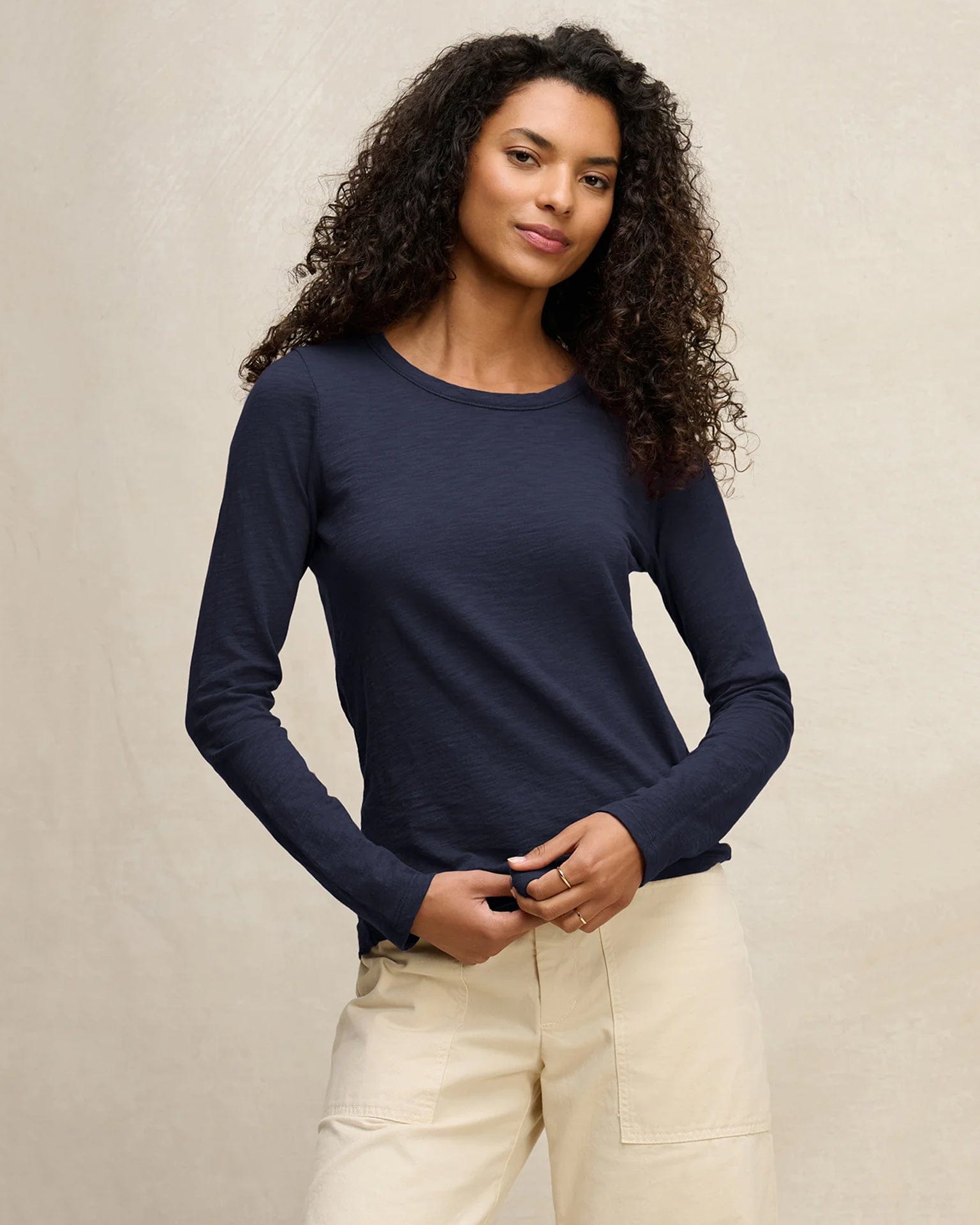 A woman with curly hair wears the Velvet by Graham & Spencer Lizzie Long Sleeve Crew in Midnight and light beige pants, standing against a neutral background, looking confidently at the camera with a slight smile.