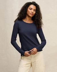 A woman with curly hair wears the Velvet by Graham & Spencer Lizzie Long Sleeve Crew in Midnight and light beige pants, standing against a neutral background, looking confidently at the camera with a slight smile.
