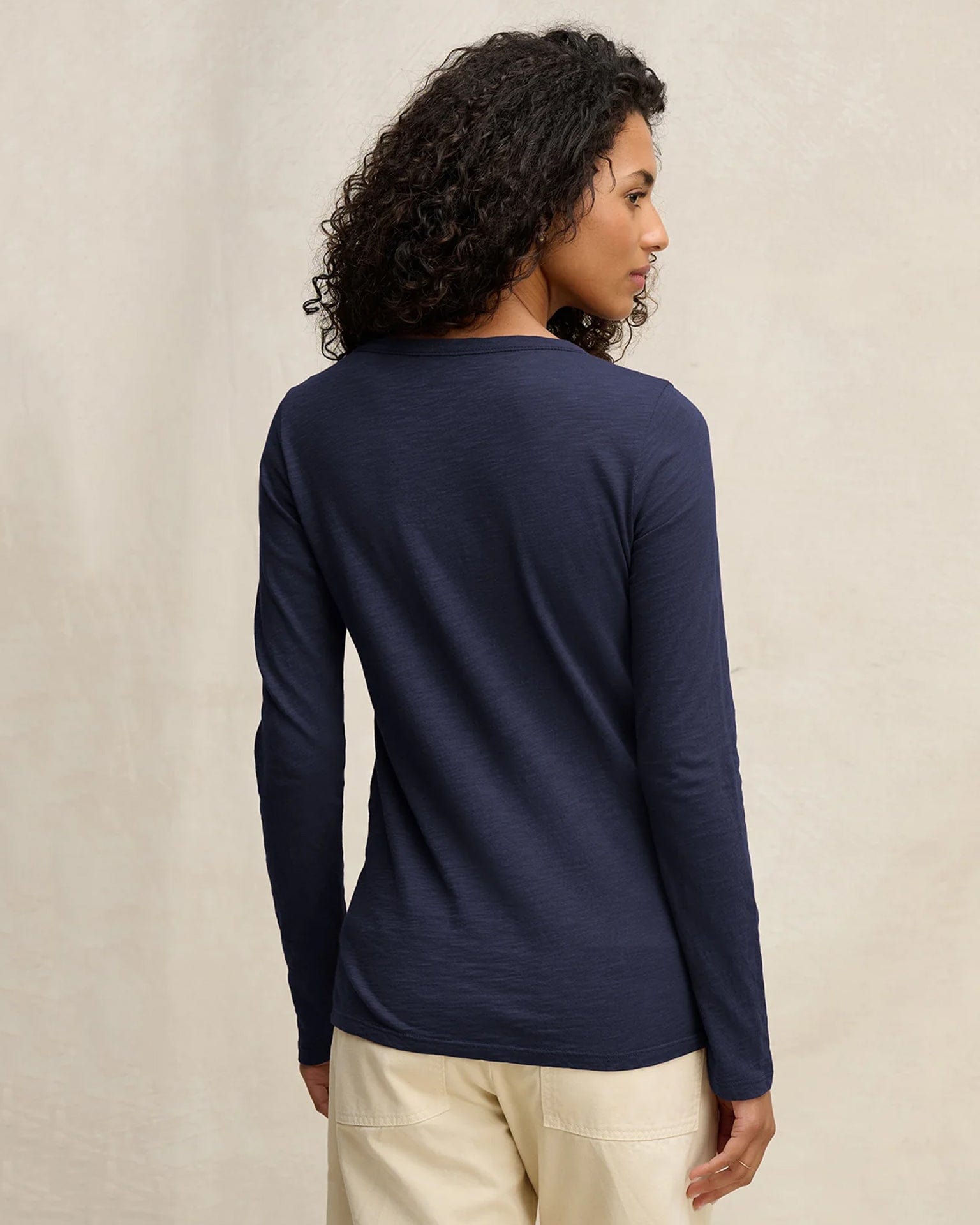 A person with curly hair, facing away from the camera against a neutral background, wears the Velvet by Graham & Spencer Lizzie Long Sleeve Crew in Midnight and light-colored pants.
