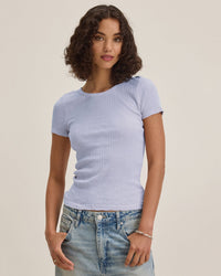 A woman with curly brown hair wears the Enati S/S Crew Neck Top in Ceramic by Velvet by Graham & Spencer and faded blue jeans. She stands against a light background, looking at the camera with a relaxed expression.