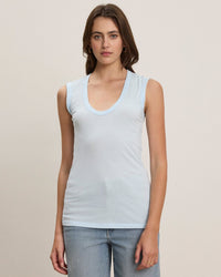 A woman with long brown hair wears a light blue Estina Scoop Neck Tank Top in Capri by Velvet by Graham & Spencer and light blue jeans, standing against a plain background and looking directly at the camera.
