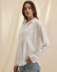 A person with long brown hair wears the Grace L/S Button Up Top in White by Velvet by Graham & Spencer and light blue jeans, standing against a cream-colored fabric backdrop.