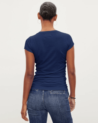 A person with short, dark hair in a neat bun is seen from behind, wearing the Velvet by Graham & Spencer Jemma S/S Crew Neck Top in Midnight, blue jeans, hoop earrings, and a bracelet against a plain light background.