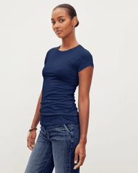 A woman stands against a plain light background, wearing the Velvet by Graham & Spencer Jemma S/S Crew Neck Top in Midnight and blue jeans. She faces slightly to the side, looking at the camera with a relaxed, confident expression.