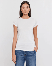 A woman with straight dark hair wears the Velvet by Graham & Spencer Jemma S/S Crew Neck Top in white and blue jeans, standing against a plain light background, looking at the camera with one hand in her pocket.