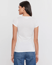 A woman with long dark hair stands with her back to the camera, wearing the Jemma S/S Crew Neck Top in White by Velvet by Graham & Spencer and blue jeans against a plain light background.