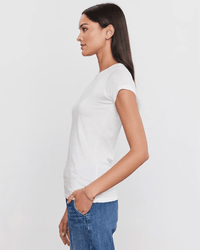 A woman with long dark hair stands in profile against a white background, wearing the Jemma S/S Crew Neck Top in White by Velvet by Graham & Spencer and blue jeans, her left hand tucked into her pocket.