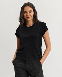 A woman with wavy dark hair wears the Marie S/S Crop Top in Black by Velvet by Graham & Spencer and black pants, standing against a light background. She smiles softly with her hands in her pockets.