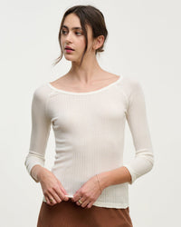 A woman with dark hair in a loose updo models the Nima L/S Boatneck Top in Ecru by Velvet by Graham & Spencer, a slim-fit, ribbed cream long-sleeve with a boat neckline. She stands against a plain background, looking to her left.
