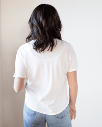 A person with shoulder-length dark hair, wearing the Velvet by Graham & Spencer Tera S/S Tee in White and light blue jeans, stands facing away from the camera against a plain light background.