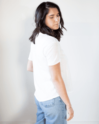 A woman with shoulder-length dark hair, wearing the Tera S/S Tee in White by Velvet by Graham & Spencer and light blue jeans, stands sideways against a plain light background, looking down with a neutral expression.