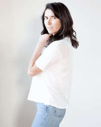 A person with shoulder-length dark hair, wearing the Velvet by Graham & Spencer Tera S/S Tee in White and light blue jeans, stands sideways and looks at the camera against a plain light background.