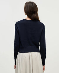 A person with long brown hair, wearing the Velvet by Graham & Spencer Violette Long Sleeve Cardi in Navy and a light skirt, stands facing away from the camera against a plain, light background.