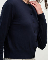 Wearing the Violette Long Sleeve Cardi in Navy by Velvet by Graham & Spencer and light pants, a person raises their hand near their face to display a gold ring. The head is partially cropped out of the frame.