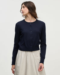 A person with long brown hair, wearing the Violette Long Sleeve Cardi in Navy by Velvet by Graham & Spencer and a light beige skirt, stands against a plain white background looking to the side.