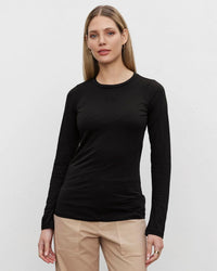 A woman with straight, shoulder-length blonde hair models the Zofina in Black top by Velvet by Graham & Spencer, featuring a fitted crew neckline and long sleeves, paired with beige pants against a plain light gray background.