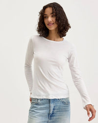 A person with curly brown hair wears Velvet by Graham & Spencer's Zofina in White, a crew neckline long sleeve pullover, and light blue jeans, standing against a plain white background and smiling slightly while looking to the side.