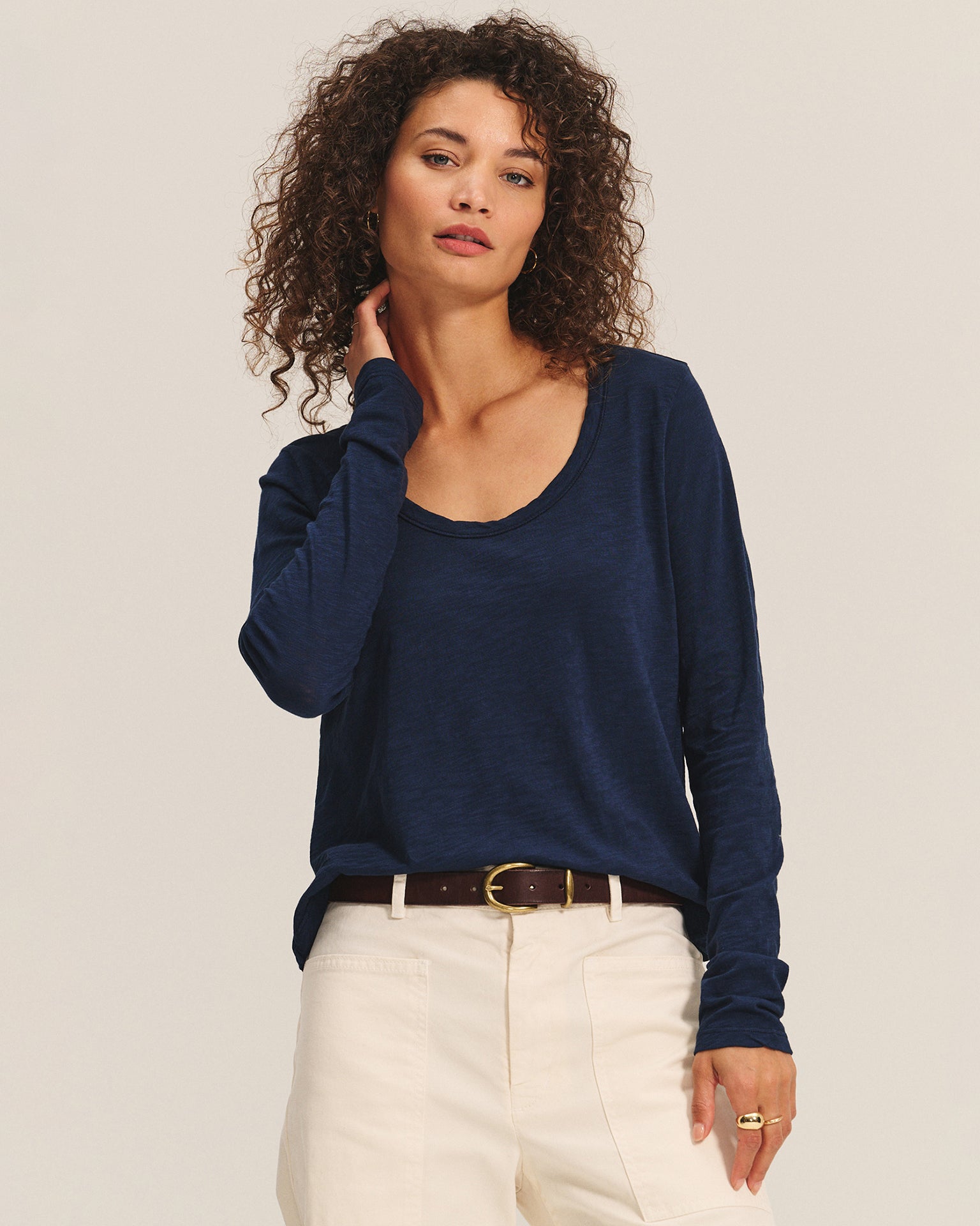 A woman with curly hair wears the Velvet by Graham & Spencer Becky Long Sleeve Scoop Neck Top in Midnight, styled with light beige pants and a brown belt, posing against a plain background with one hand touching her neck.