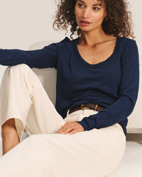 A woman with curly hair sits on a light chair, wearing the Velvet by Graham & Spencer Becky Long Sleeve Scoop Neck Top in Midnight with cream wide-leg pants, gold hoop earrings, and a dark brown belt, looking slightly to the side.