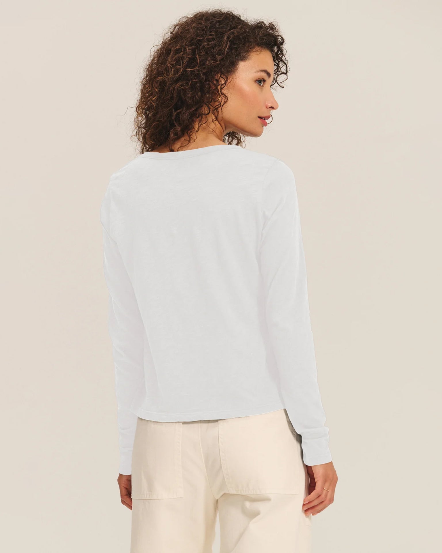 A woman with curly hair stands facing away, wearing the Velvet by Graham & Spencer Becky Long Sleeve Scoop Neck Top in White and cream pants against a plain light background.