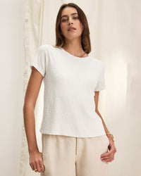 A woman wearing a Velvet by Graham & Spencer Marika Short Sleeve Crew Neck Crop Top in white and light beige pants stands against a cream-colored fabric backdrop, looking at the camera with a relaxed expression.