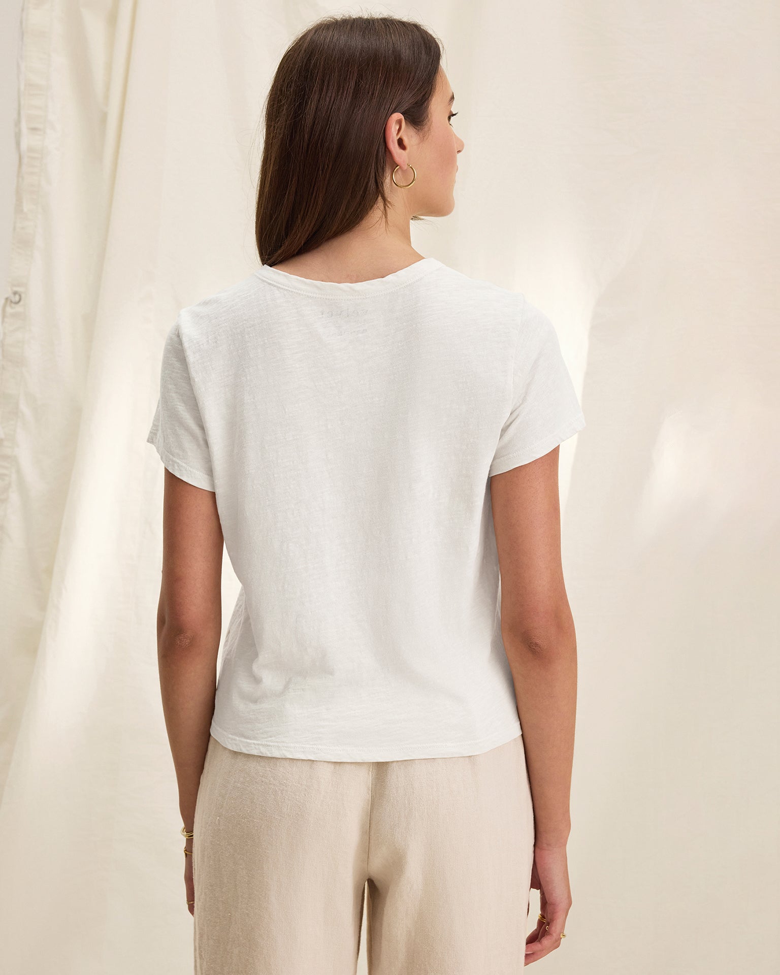 A woman with straight brown hair stands facing away, wearing a Velvet by Graham & Spencer Marika Short Sleeve Crew Neck Crop Top in white and light beige pants against a soft, cream-colored fabric backdrop.