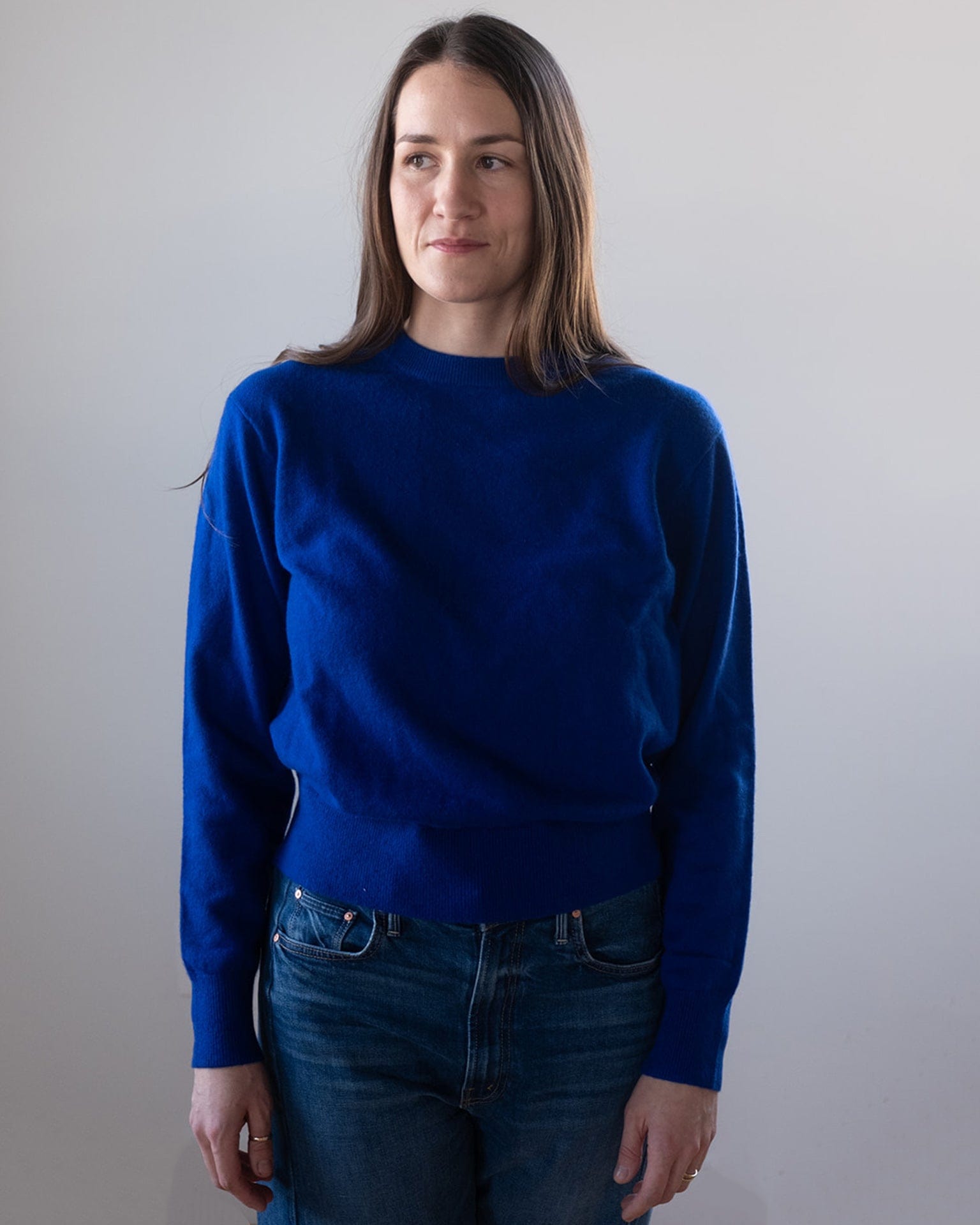 A woman with long brown hair wears the White & Warren Cashmere Crew in Frontier Blue with blue jeans, standing against a plain light-colored background and looking slightly to her left.